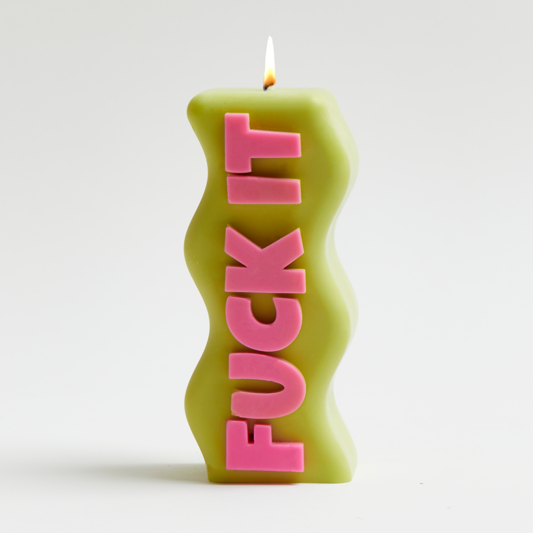 A green pillar candle with the words 'FUCK IT' in pink letters on it, with a lit flame at the top.