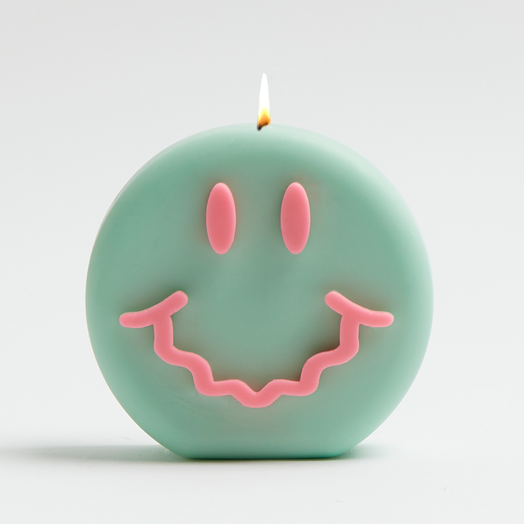 A round candle with a smiling face design in pink on a mint green background