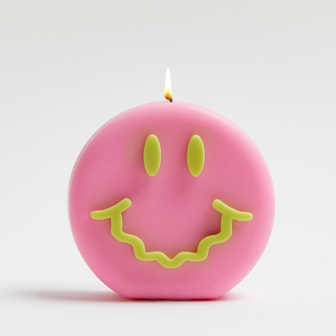A pink candle with a smiling face design, featuring green eyes and mouth.