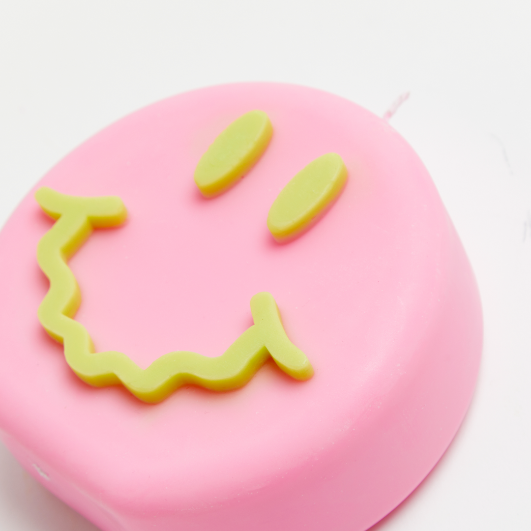 A pink candle with a smiling face design, with green eyes and mouth.