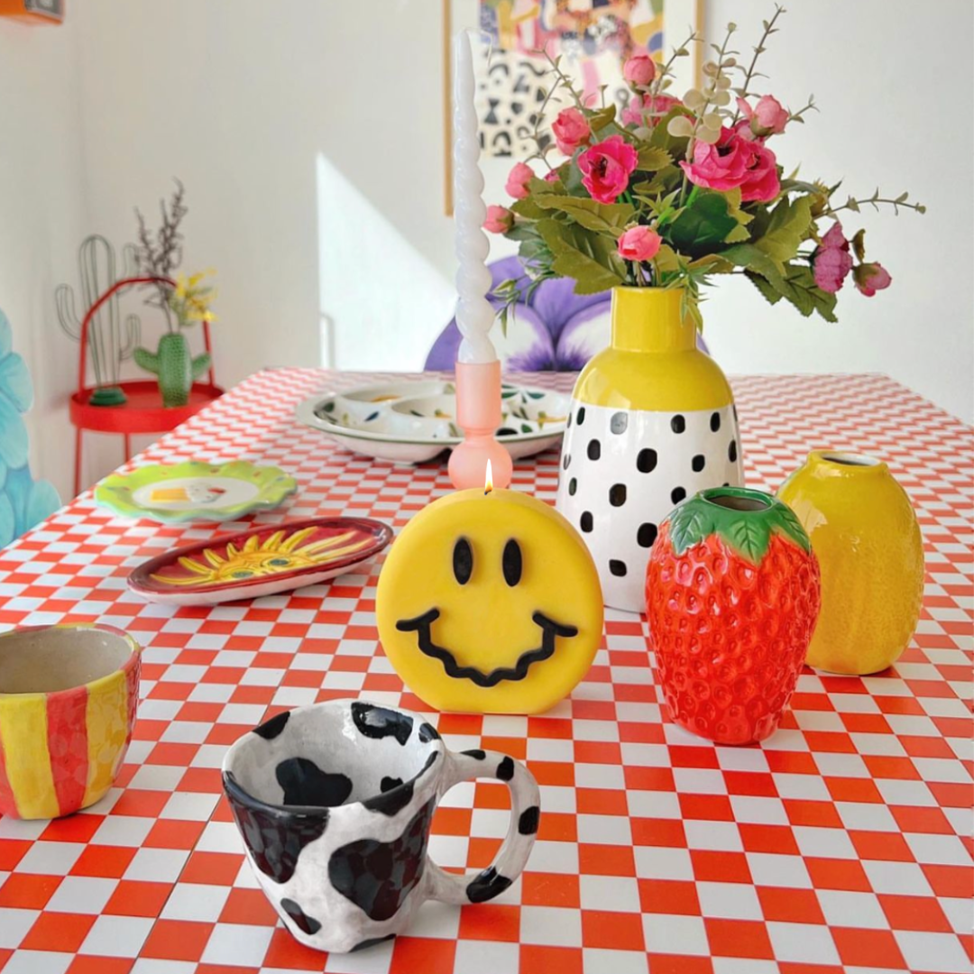 Yellow round sculptural candle with pink smiling face design and lit wick on a table
