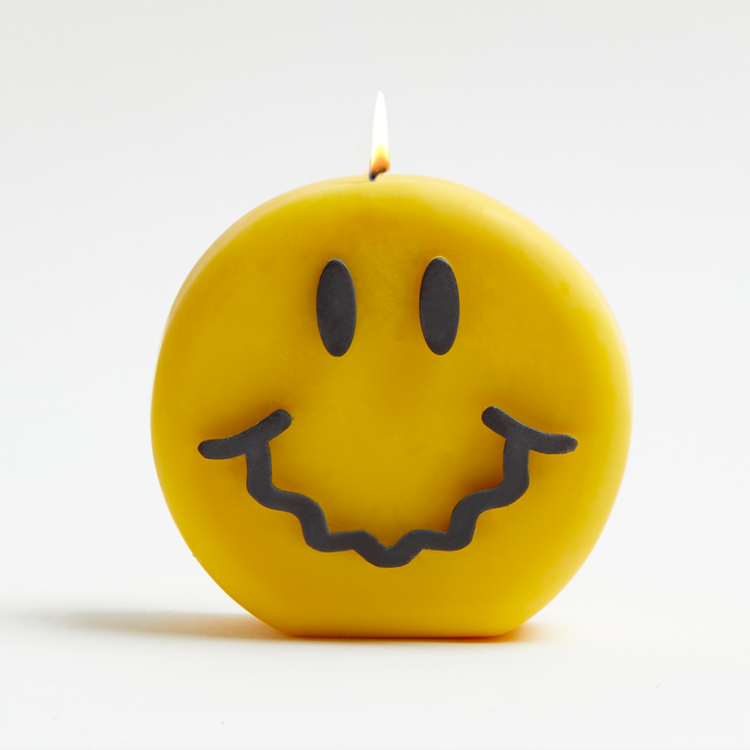 A yellow candle with a black smiling face design and a lit flame, on a white background.