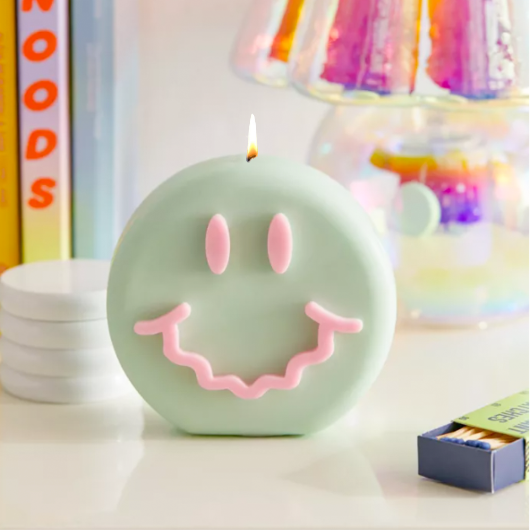 Green round sculptural candle with pink happy face design and lit wick