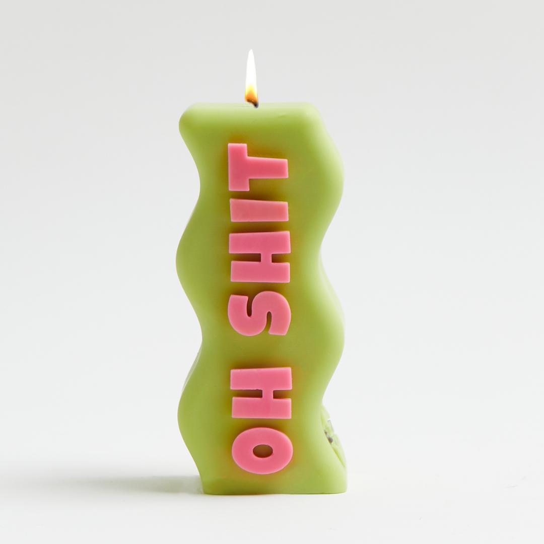 A green candle with pink text spelling 'OH SHIT' on it, with a flame at the top.
