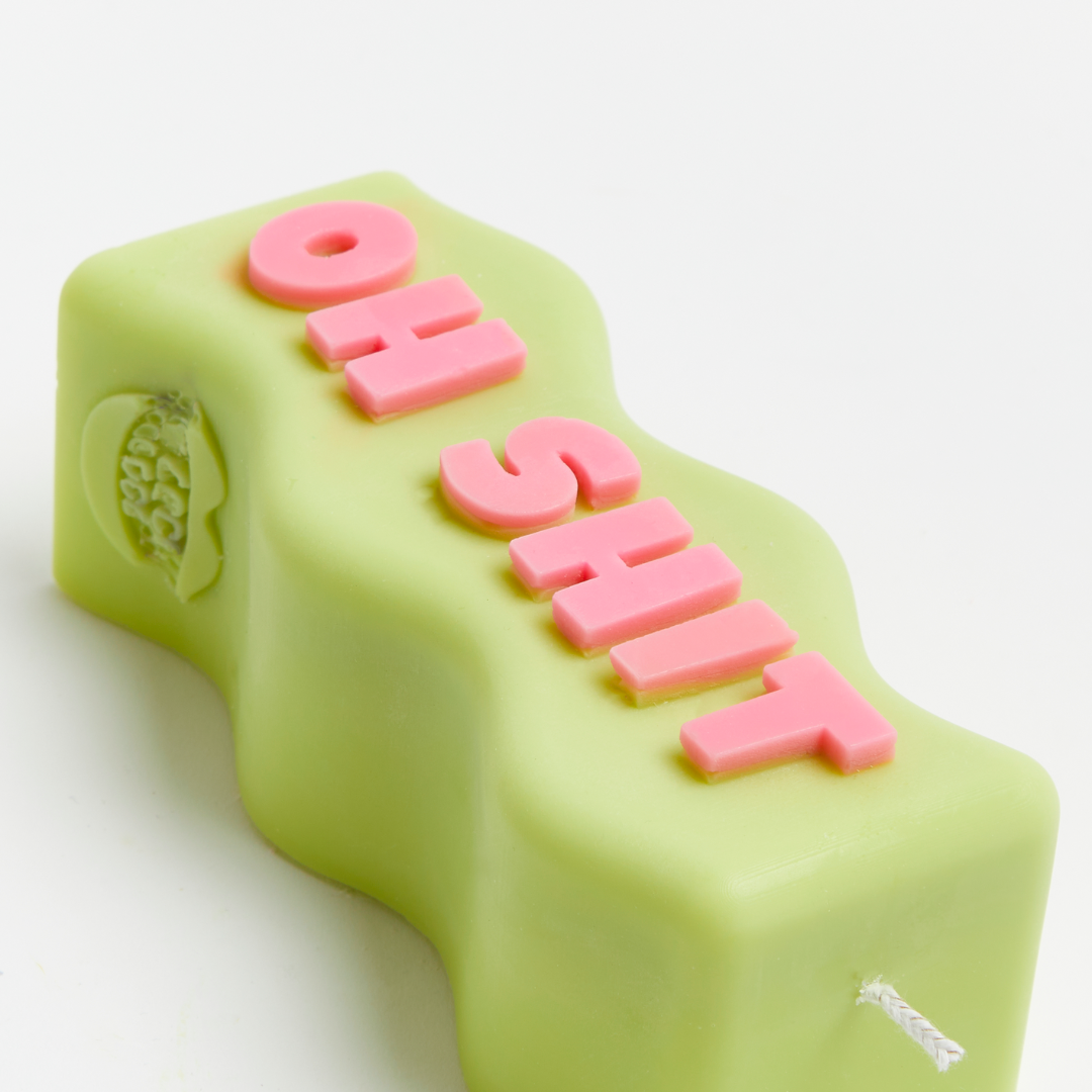 A green candle with pink text spelling 'OH SHIT' on it, with a flame at the top.