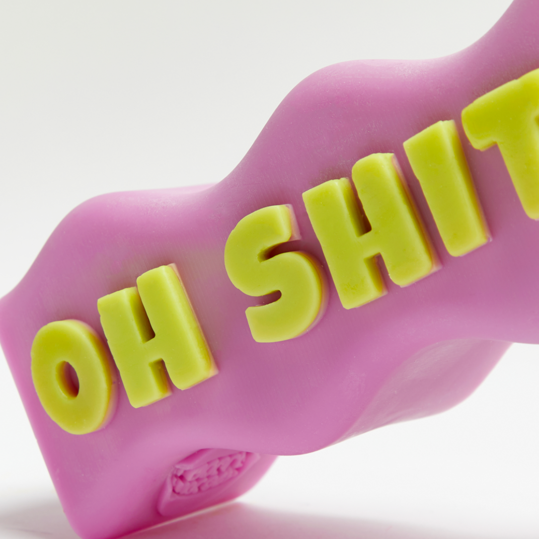 Purple sculptural statement candle spelling OH SHIT with green lettering and lit wick