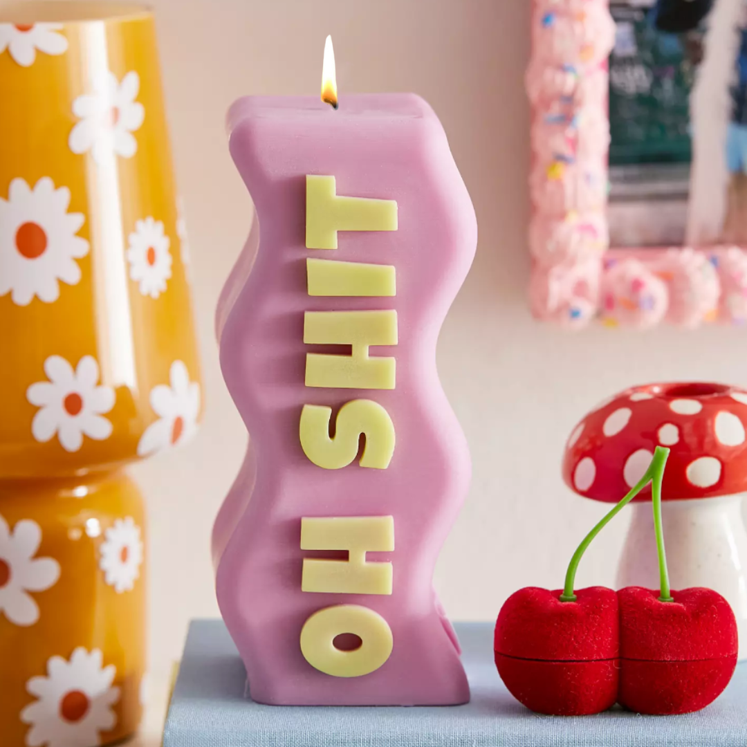 Purple sculptural statement candle spelling OH SHIT with green lettering and lit wick